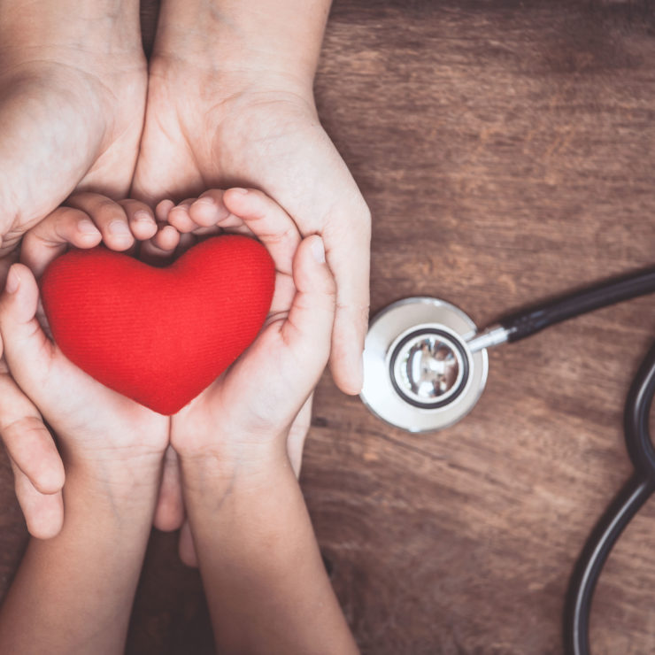 Woman and child with their hands lying on top of each other holding a knitted red heart with a doctor's stethoscope in the background showing how integrating compassion into medical care is the necessary next step for the field.